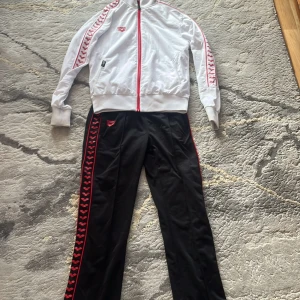 Svarta träningsbyxor från Arena - Svarta träningsbyxor från Arena med röda detaljer och logga längs sidorna. Byxorna har en rak passform och är tillverkade i ett glansigt syntetmaterial som känns sportigt och bekvämt. Perfekta för träning eller chill dagar.