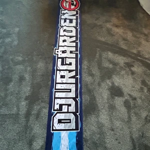 Djurgården supporterhalsduk blåvit - Snygg lång supporterhalsduk med texten 'DJURGÅRDEN' på ena sidan och 'FRIHET FÖR ULTRA' på andra. Halsduken har blå och vita färger med stora, tydliga bokstäver och en grafisk detalj upptill. Perfekt för dig som vill visa ditt stöd på läktaren.