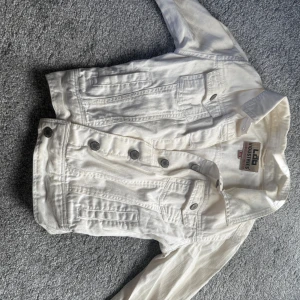 Vit jeansjacka med spets från Lab Industries stl. XXS - Snygg vit jeansjacka från Lab Industries med coola silverfärgade knappar och broderad spetsdetalj på ryggen. Klassisk krage, långa ärmar och flera fickor framtill. Perfekt för dig som vill ha en unik twist på en klassisk jacka.