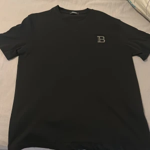 Svart t-shirt från Balmain med B-logga - Snygg svart t-shirt från Balmain med en stilren silverfärgad B-logga på bröstet. Klassisk rund hals och korta ärmar. Materialet är mjukt och känns lyxigt mot huden, perfekt för dig som gillar exklusiva märken och enkel design.