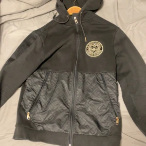 Svart hoodie från Glorious Gangsta - Svart hoodie från Glorious Gangsta med guldfärgade detaljer och dragkedja. Nedre delen har ett diskret mönster och två stora fickor med dragkedja. Märkets logga i guld på bröstet och huva med snörning ger en riktigt clean streetlook.