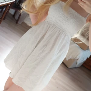 Vit axelbandslös kortklänning - Supersöt vit axelbandslös kortklänning  med broderade blommor och smock upptill. Klänningen har en liten guldfärgad logoknapp nertill och är gjord i bomull, perfekt för varma dagar.