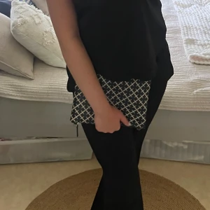 Svartvit rutig kuvertväska - Snygg kuvertväska i svart med vita rutmönster, perfekt att bära i handen. Väskan har en rektangulär form och stängs med dragkedja. Stilren design som passar till många outfits och är enkel att matcha.