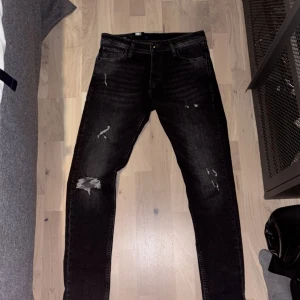 Svarta slitna jeans från Jack & Jones - Svarta jeans från Jack & Jones med slitningar och trasiga detaljer på benen. Modellen är slim fit med klassisk femficksdesign och knappgylf. W 30 L 32 