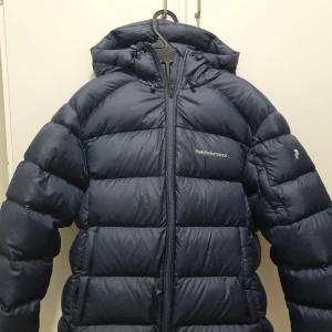 Mörkblå dunjacka Peak Performance - Snygg och varm dunjacka från Peak Performance i storlek M. Fint marinblå färg som passar till det mesta. Jackan är använd få gånger och i mycket gott skick – inga skador, fläckar eller slitage. Perfekt för kalla vinterdagar tack vare den tjocka dunfyllningen och den sköna huvan. 