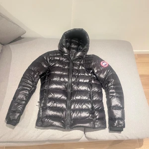 Svart dunjacka från Canada Goose - Säljer en svart dunjacka från Canada Goose med huva och klassisk logga på ärmen. Jackan är quiltad, har dragkedja framtill och två sidofickor med dragkedja. Perfekt för kalla dagar och har en riktigt snygg, puffig look.