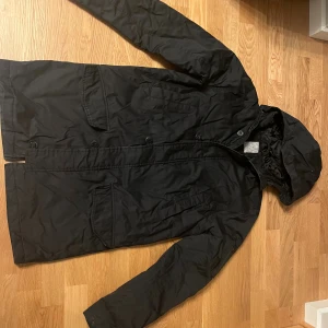 Svart Etirel parkajacka med huva - Svart Etirel parkajacka med huva och flera fickor framtill. Jackan har lång passform och stängs med knappar och dragkedja. Perfekt för kyligare dagar och har en stilren look som funkar till det mesta.