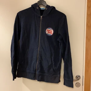 Mörkblå hoodie från Jack & Jones - Säljer en mörkblå hoodie från Jack & Jones Originals i storlek medium. Tröjan har dragkedja, huva med snörning och en snygg logga på bröstet. Perfekt för chill dagar och enkel att matcha med jeans eller joggers. Lite färgblekt o ett pyttehål vid fickan som jag har sytt igen. 