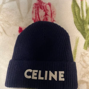 Mörkblå ribbad mössa från Celine - Snygg mörkblå ribbstickad mössa från Celine med uppvikt kant och stor vit logga framtill. Perfekt accessoar för att hålla värmen och ge din outfit en lyxig touch. Klassisk form och stilren design som passar till det mesta.