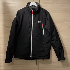 Svart vindjacka från Helly Hansen M - Svart vindjacka från Helly Hansen med vita och röda detaljer. Jackan har hög krage, flera dragkedjeförsedda fickor och HH-logga på bröstet och ryggen. Perfekt för blåsiga dagar och har en sportig look med snygg passform.