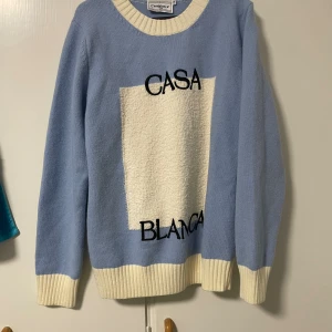  Casablanca tröja - Snygg ljusblå stickad tröja från Casablanca med kontrasterande vita ribbade muddar och krage. Endast testad annat helt ny. Framsidan har ett vitt block och svart text med 'CASABLANCA'. Perfekt för dig som gillar stilrena och trendiga plagg med en sportig touch.