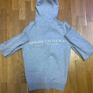 Armani Hoodie - Säljer en stilren grå hoodie från Armani Exchange med logga tryckt på bröstet och stor text på ryggen. Tröjan har huva med snörning och är långärmad. Perfekt för dig som gillar en clean och modern look.