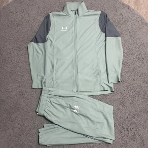 Under Armour träningsset grå/grön - Snyggt träningsset från Under Armour i ljusgrå/grön färg med mörkgrå detaljer på axlarna. Jackan har dragkedja framtill och byxorna har resår och snörning i midjan samt dragkedjor vid bensluten. Perfekt för träning eller chill.