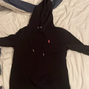 Svart hoodie från Polo Ralph Lauren - Svart hoodie från Polo Ralph Lauren med klassisk rosa broderad logga på bröstet. Tröjan har huva med snörning, magficka och ribbade muddar. Perfekt för en chill och stilren look.