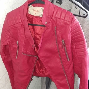 Röd skinnjacka från Vogelle, stl 36 - Snygg röd skinnjacka från Vogelle i bikerstil med coola quiltade axlar och detaljerade dragkedjor framtill. Jackan har en figurnära passform och långa ärmar, perfekt för dig som vill sticka ut med en färgstark look.