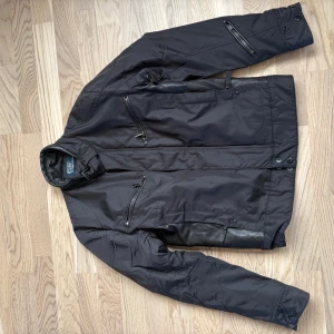 Svart jacka Polo Ralph Lauren S - Svart jacka från Polo Ralph Lauren i storlek S. Jackan har flera dragkedjor, hög krage med spänne och detaljer i skinn. Materialmix av polyester och skinn ger en cool look. Perfekt för höst och vinter, med lång ärm och snygg passform.