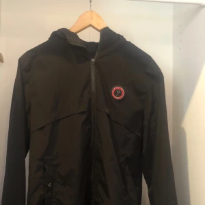 Svart vindjacka från Trapstar med huva - Svart vindjacka från Trapstar med huva och dragkedja. Stor broderad Trapstar-logga i rött på ryggen och rund patch på bröstet. Jackan har långa ärmar och praktiska fickor framtill. Perfekt för dig som vill ha streetwear-vibe.