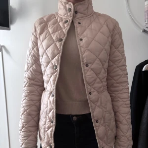 Beige quiltad jacka med tryckknappar - Säljer en beige quiltad jacka med hög krage och tryckknappar framtill. Jackan har ett rutigt mönster, två fickor och en figurnära passform. Modellen brukar bära xs/s. Skriv privat om ni har några frågor.