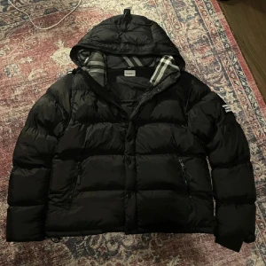 Svart dunjacka från Burberry - Säljer en svart dunjacka från Burberry med klassiskt rutigt foder i huvan. Jackan har dragkedja, knappar och två fickor framtill. Märkespatch på ärmen och en skön, puffig känsla som håller dig varm hela vintern.