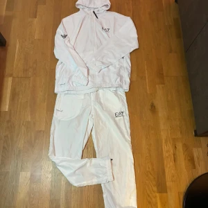 Vit vindjacka från EA7 Emporio Armani - Säljer en fräsch vit vindjacka från EA7 Emporio Armani med huva och hel dragkedja. Jackan har logga på bröstet och ärmen, elastiska muddar och lätt material som passar perfekt för chill och sportiga dagar. Snygg och stilren design med modern vibe.