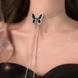 Silverfärgat halsband med svart fjäril - Snyggt halsband i silverfärg med en svart fjäril som hängsmycke. Kedjan är tunn och har två långa hängen som ger en cool och modern look. Fjärilen har glittriga detaljer runt kanterna som gör att den sticker ut. Perfekt accessoar för att lyfta din outfit.