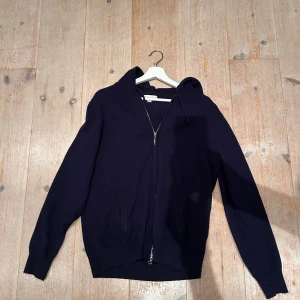 Mörkblå hoodie från Barluccio - Mörkblå hoodie från Barluccio med dragkedja framtill och två fickor. Tröjan är stickad och har en klassisk huva med dragsko. Perfekt för dig som gillar en enkel och stilren look med lite sportig känsla.