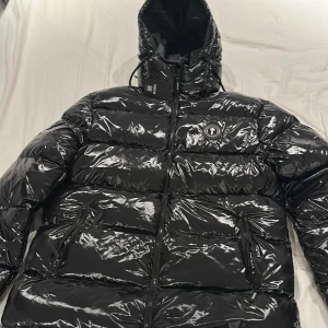 Svart pufferjacka från Trapstar - Snygg svart pufferjacka från Trapstar med glansig finish och tydlig logga på bröstet och ryggen. Jackan har huva, dragkedja framtill och två fickor med dragkedja. Perfekt för kalla dagar när du vill sticka ut med streetstyle.