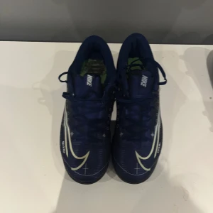 Blå Nike fotbollsskor MDS - Säljer ett par blå Nike fotbollsskor med vita och ljusblå detaljer samt MDS-tryck på sidan. Skorna har snörning, platt sula och är designade för inomhusspel. Swoosh-loggan syns tydligt på både ovansida och sula. Perfekta för dig som vill sticka ut på planen.