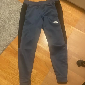 Blå träningsbyxor från The North Face - Snygga blå träningsbyxor från The North Face med svarta paneler längs sidorna. Byxorna har en smal passform, elastisk midja och logga på låret. Perfekta för träning eller chill. Tillverkade i mjukt och stretchigt syntetmaterial.