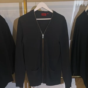 Svart kofta med dragkedja från Hugo Boss - Stilren mörk grå kofta från Hugo Boss med dragkedja framtill och två fickor. Tillverkad i en skön mix av bomull och ull, perfekt för lager-på-lager. Klassisk design med lång ärm och diskret kabelstickad detalj längs dragkedjan.