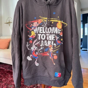Space Jam svart hoodie med tryck Siksilk - Svart hoodie med stort Space Jam-tryck framtill och texten 'Welcome to the Jam!'. Färgglada Looney Tunes-karaktärer och basketmotiv. Huva med snörning och känguruficka. Perfekt för dig som älskar streetwear och nostalgi.