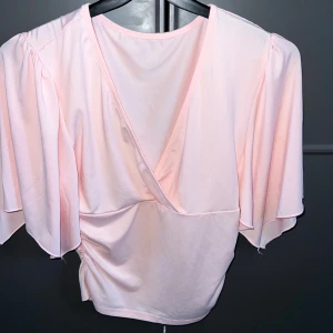 Ljusrosa omlottopp från SHEIN - Säljer en ljusrosa omlottopp från SHEIN i storlek S. Toppen har v-ringning, korta och vida ärmar samt snygg rynkning på sidan. Materialet är mjukt och stretchigt, perfekt för dig som gillar en feminin och trendig look.