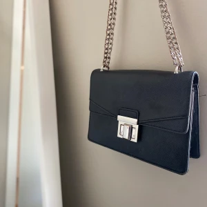 Svart axelväska med kedja - Stilren svart axelväska med rektangulär form och grov silverfärgad kedja som axelrem. Väskan har ett snyggt metallspänne framtill och är tillverkad i skinnliknande material. Insidan har ett fack med dragkedja för småsaker. Perfekt för dig som gillar minimalistisk design . Stor rymlighet och passar till allt en av de bästa väskorna jag har ägt då man får plats med sjukt mycket grejer och mer än man tror jätte bra faktiskt ❣️ köpt från Zara 