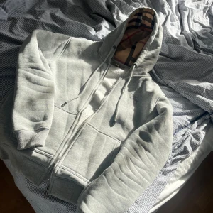 Grå hoodie från Burberry med dragkedja - Säljer en grå hoodie från Burberry i 100% bomull med klassiskt rutigt foder i huvan och arm siddorna. Tröjan har dragkedja framtill, snörning i huvan och fickor Perfekt för dig som vill ha en stilren och lyxig still med ikoniska Burberry-detaljer. 