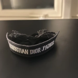 Christian Dior J'Adior armband grått - Snyggt svart och grått armband från Christian Dior med texten 'J'Adior' broderad i vitt. Armbandet har flätade snören och tofsar i ändarna för justerbar passform. Perfekt accessoar för dig som gillar lyxiga detaljer och statement pieces.