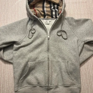 Grå Burberry hoodie med dragkedja - Snygg grå hoodie från Burberry med klassiskt rutigt foder i huvan och dragkedja framtill. Tröjan har snörning i huvan, känguruficka och är tillverkad i mjuk bomull. Perfekt för dig som vill ha en stilren och lyxig vardagslook. Storlek M men sitter mer som S-Xs
