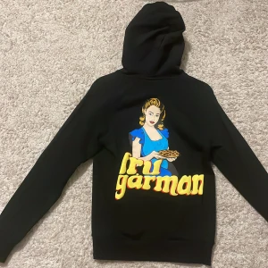 Svart hoodie Fru Garman Revenite XS - Svart hoodie från Revenite i storlek XS med stor färgglad print på ryggen av en kvinna i blå klänning och texten 'fru garman' i gul 3D-stil. Klassisk huva och långa ärmar, perfekt för dig som gillar unika tryck och streetstyle.