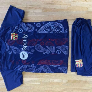 FC Barcelona Lamine Yamal fotbollströja - Säljer en blå FC Barcelona fotbollströja med Lamine Yamal och nummer 19 på ryggen. Tröjan har korta ärmar, paisleymönster i vitt och rött, samt klubbmärke och Spotify-logga framtill. Tillverkad i lätt och ventilerande syntetmaterial. Matchande shorts medföljer.