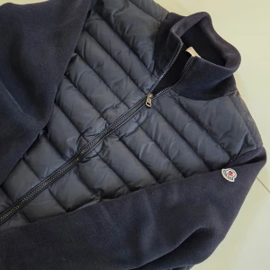 Mörkblå quiltad jacka från Moncler - Snygg jacka från Moncler med quiltad front och stickade ärmar och rygg. Jackan har dragkedja framtill, ståkrage och två fickor med dragkedja. Klassisk Moncler-logga på ärmen. Perfekt för dig som vill ha en stilren och sportig look.