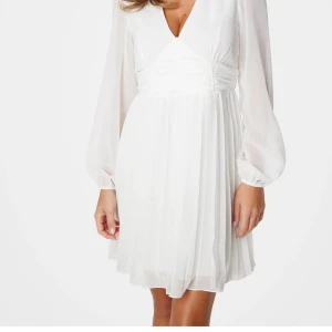 Vit plisserad kortklänning med v-ringning - Supersnygg vit kortklänning med plisserad kjol och v-ringad hals. Klänningen har långa, transparenta ärmar och markerad midja med rynkade detaljer. Tillverkad i ett lätt och luftigt material som ger en elegant känsla. Perfekt för dig som vill ha en fräsch och stilren look. Endast använt 3 gånger! Ny pris: 420kr