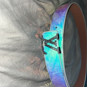 Louis Vuitton holografiskt bälte - Säljer ett Louis Vuitton bälte med bred holografisk rem i blå, lila och turkosa toner. Bältet har en stor LV-logga i svart med glittriga detaljer och klassiskt monogrammönster. Insidan är i ljust brunt skinn med präglad märkning. Perfekt statement piece för din outfit.
