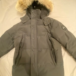 Svart dunjacka från Canada Goose - Säljer en svart dunjacka från Canada Goose med klassisk patch på ärmen och stor huva med beige fuskpäls. Jackan är quiltad, har dragkedja och knappar framtill samt två stora fickor. Perfekt för kalla vinterdagar och riktigt snygg streetstil. Inget kvitto 