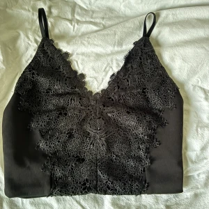 Svart spetslinnetopp med smala band - Snygg svart linnetopp med tunna axelband och detaljerad spets framtill. Toppen har v-ringning och croppad passform.