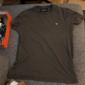 Mörkbrun t-shirt från Lyle & Scott - Säljer en mörkbrun t-shirt från Lyle & Scott i storlek medium. T-shirten har klassisk rund hals, korta ärmar och är gjord i mjuk bomull. På bröstet sitter den ikoniska gula fågeln broderad. Perfekt för dig som gillar stilrena och enkla plagg.