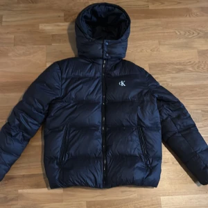 Svart pufferjacka från Calvin Klein Jeans, Storlek S - Snygg svart pufferjacka från Calvin Klein Jeans med huva och dragkedja framtill. Jackan har två fickor med dragkedja och diskret cK-logga på bröstet. Perfekt för kalla dagar och riktigt stilren look i storlek S.Ny pris 2700kr