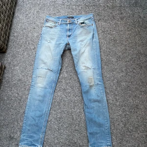 Nudie jeans - Säljer ett par ljusblå s jeans från Nudie Jeans med slitna detaljer och trasiga partier på benen. Klassisk femficksmodell med orange sömmar och Nudie-logga på bakfickorna. 