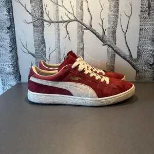 Röda/Vinröda Puma sneakers i mocka - Säljer ett par klassiska Puma sneakers i vinröd mocka med vita detaljer och snörning. Skorna har en låg profil, vit sula och den ikoniska Puma-loggan på sidan. Perfekta för dig som gillar retrostil och vill sticka ut med färg.