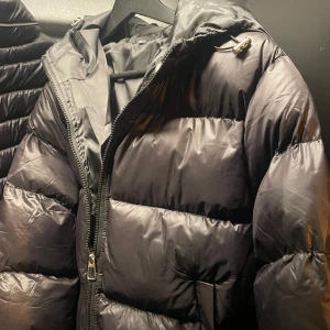 Svart pufferjacka med dragkedja - Säljer en svart pufferjacka med hög krage och huva. Jackan har en rejäl dragkedja framtill och breda, vadderade paneler som ger en riktigt fet look. Perfekt för kalla dagar när du vill hålla stilen och värmen. HELT NY!