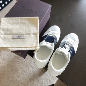 Valentino Garavani Open sneakers vit/blå - Säljer ett par snygga Valentino Garavani Open sneakers i vitt skinn med en bred, mörkblå lackad rem över vristen. Klassisk rund tå, vita snören och diskret logga på plösen. Kommer med originalkartong och dustbag. Perfekta för dig som gillar lyxiga detaljer.