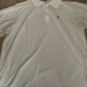 Vit piké från Ralph Lauren - Vit pikétröja från Ralph Lauren med klassisk krage och korta ärmar. Tröjan har knappar framtill och den ikoniska lilla logotypen broderad på bröstet. Perfekt för en clean och fräsch look.